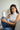 A woman sitting indoors holds a white cylindrical jar labeled 'Kris Wellness' against her knee. She is wearing a white ribbed turtleneck top and blue jeans, with a neutral indoor background and a lamp visible behind her.